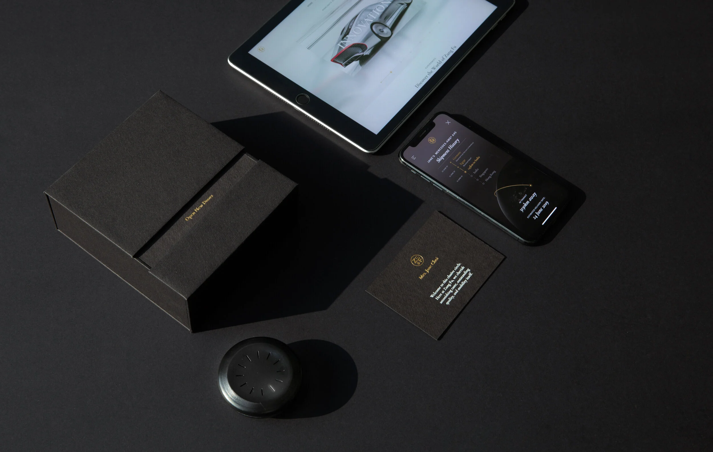 The full Open New Doors welcome kit flat-laid on black: presentation box, brand tablet, phone showing shipment history, personalised welcome card and the finished dark key.